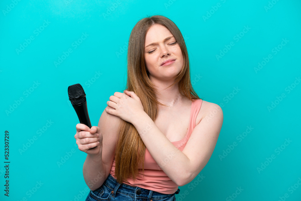 Singer caucasian woman picking up a microphone isolated on blue background suffering from pain in shoulder for having made an effort Singer caucasian woman picking up a microphone isolated on blue background suffering from pain in shoulder for having made an effort