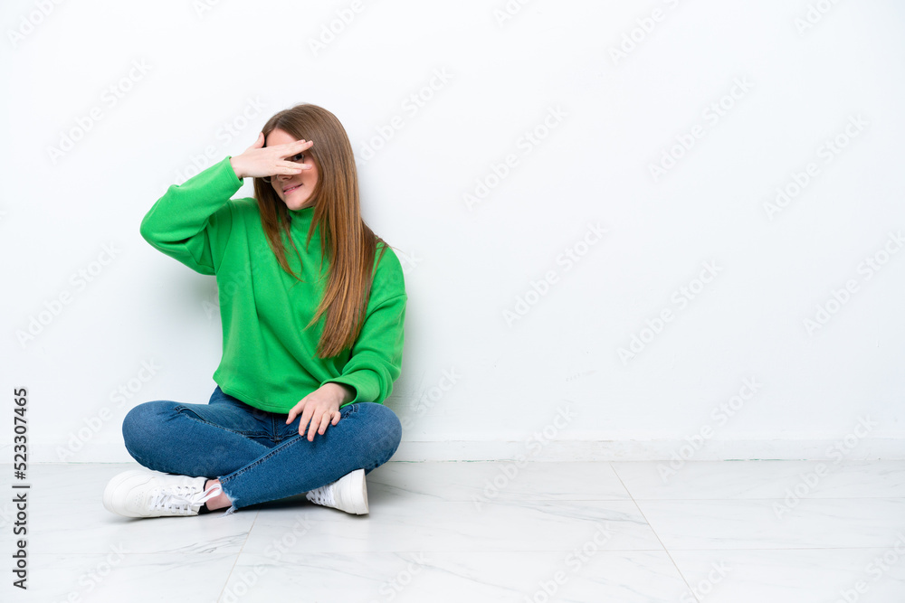 Young caucasian woman sitting on the floor isolated on white background covering eyes by hands and smiling