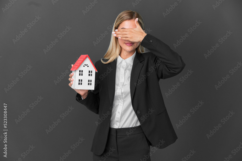 Blonde Uruguayan girl holding a house toy isolated on black background covering eyes by hands. Do not want to see something Blonde Uruguayan girl holding a house toy isolated on black background covering eyes by hands. Do not want to see something