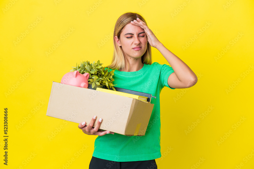 Young Uruguayan girl making a move while picking up a box full of things isolated on yellow background with headache Young Uruguayan girl making a move while picking up a box full of things isolated on yellow background with headache