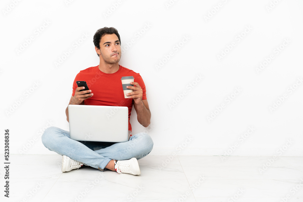 Caucasian handsome man with a laptop sitting on the floor holding coffee to take away and a mobile while thinking something Caucasian handsome man with a laptop sitting on the floor holding coffee to take away and a mobile while thinking something