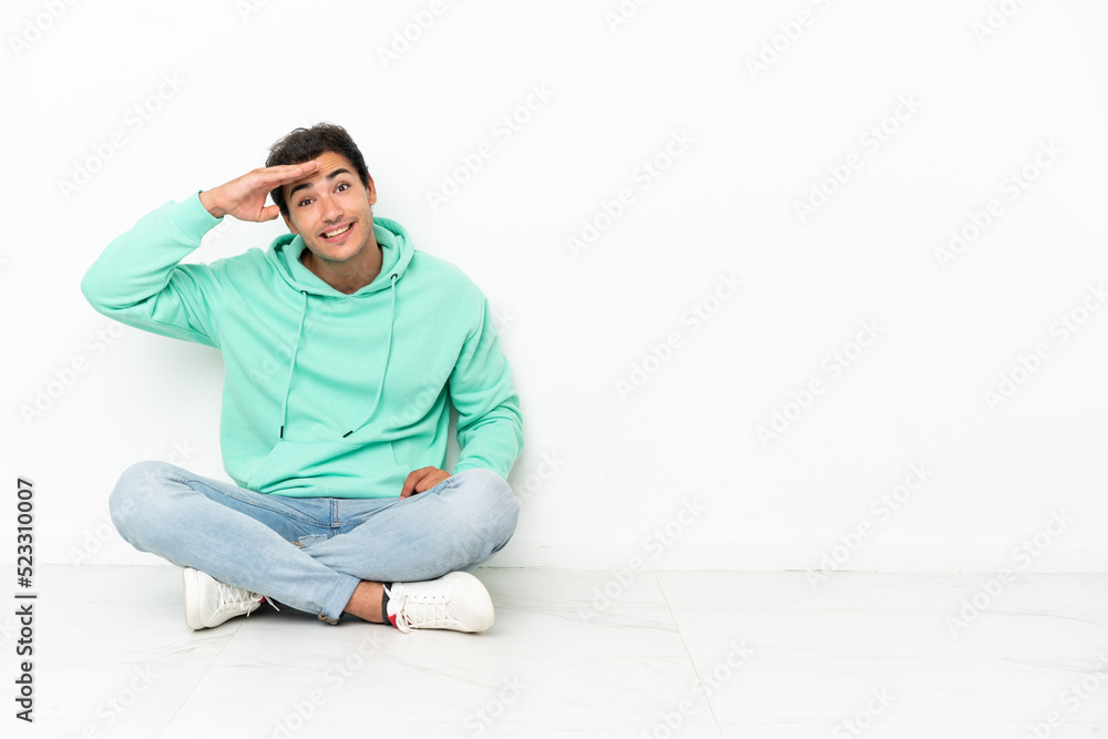 Caucasian handsome man sitting on the floor looking far away with hand to look something Caucasian handsome man sitting on the floor looking far away with hand to look something
