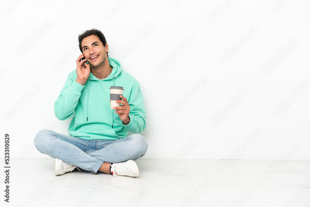 Caucasian handsome man sitting on the floor holding coffee to take away and a mobile Caucasian handsome man sitting on the floor holding coffee to take away and a mobile