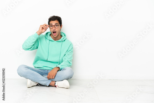 Caucasian handsome man sitting on the floor with glasses and surprised