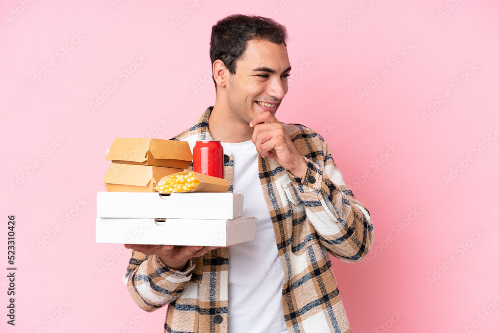 Young caucasian man holding fast food isolated on pink background looking to the side and smiling Young caucasian man holding fast food isolated on pink background looking to the side and smiling
