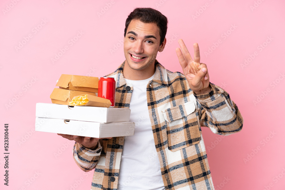 Young caucasian man holding fast food isolated on pink background happy and counting three with fingers Young caucasian man holding fast food isolated on pink background happy and counting three with fingers