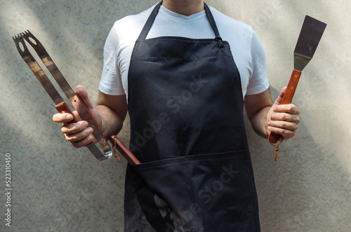 Fotografiet A man wearing black chef's apron, holding barbecue tools: bbq tongs, spatula