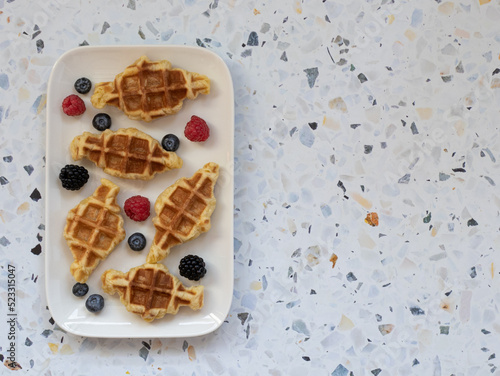 Top view of croffles (combination of croissant and belgian waffle) with fresh berries served in rectangular white dish on terrazzo background. Food trend from South Asia. Space for text