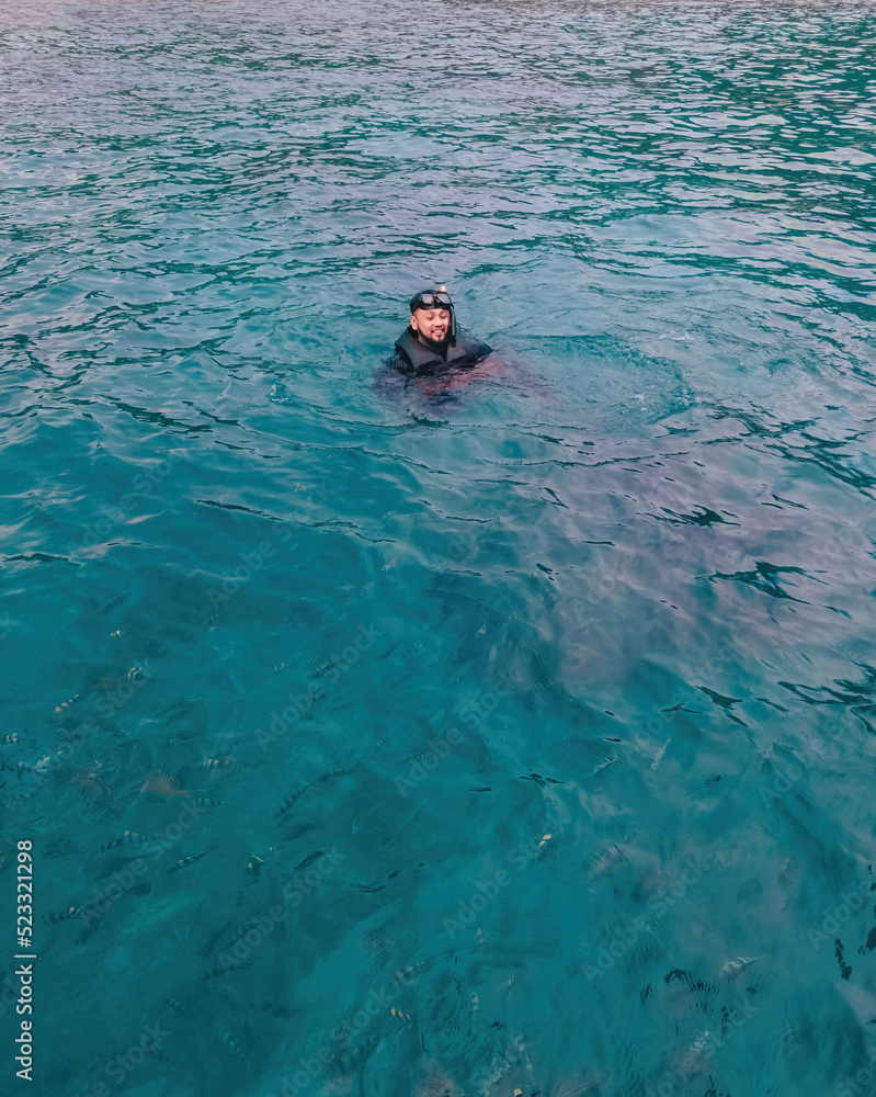 asian man wearing snorkel floating in open water surrounded by fishes.