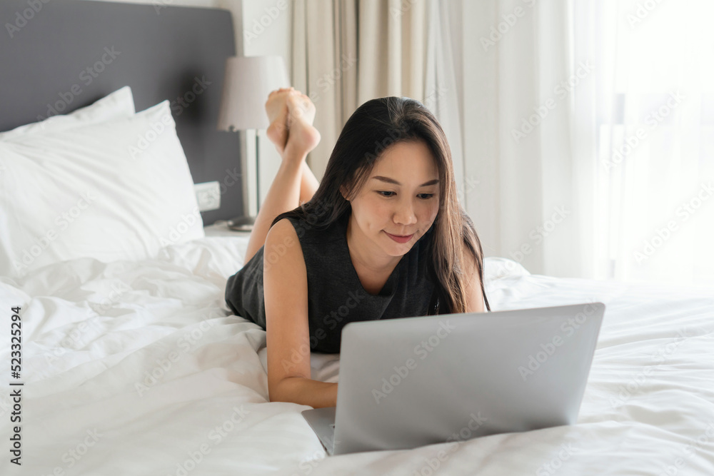 Smiling Asian businesswoman working on laptop computer in bed at hotel. Business trip, people and technology concept, copy space Smiling Asian businesswoman working on laptop computer in bed at hotel. Business trip, people and technology concept, copy space