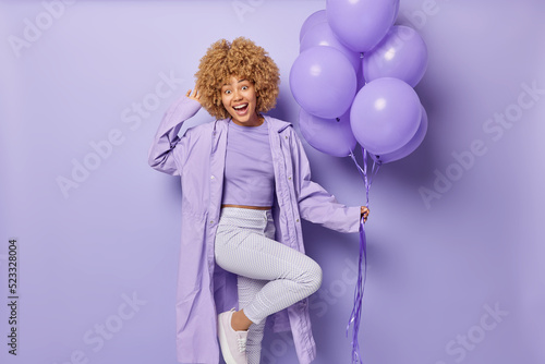 Positive glad woman stands on one leg with bunch of balloons dressed in one colored clothes comes on party to celebrate college leaving looks with cheerful expression has festive mood poses indoor