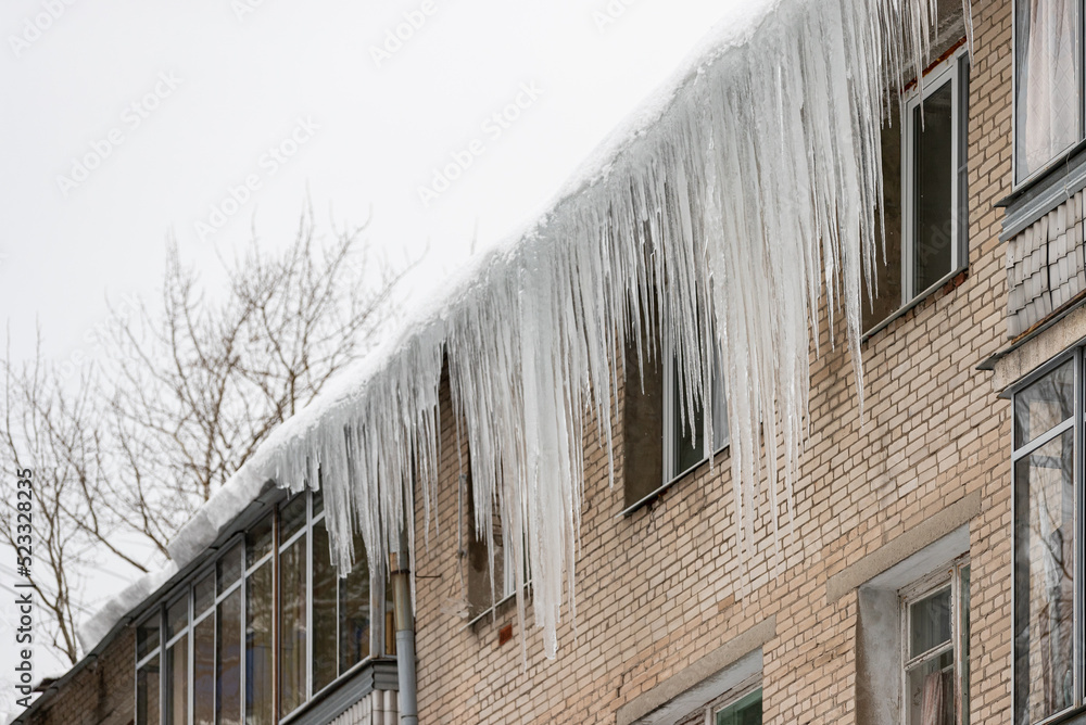 Large icicles hang from the snow-covered roof in front of the windows of the house. Snow on the roof.