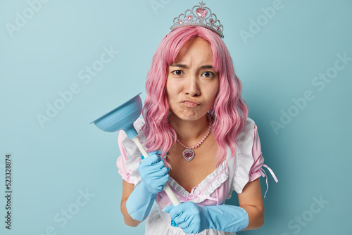 Fotografi Upset Asian woman frowns face looks displeased holds plunger ready for doing housework wears small crown rubber gloves and white dress isolated over blue background