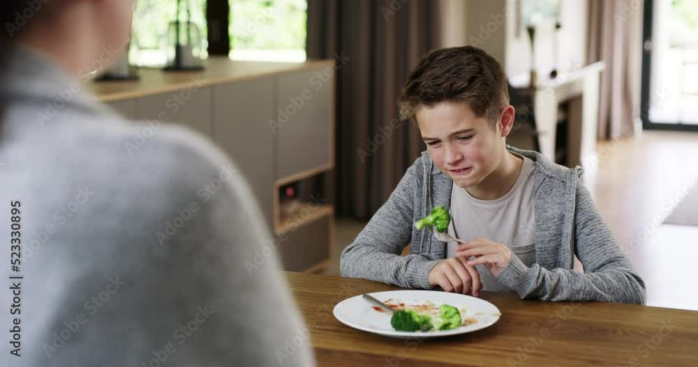 Unhappy child forced to eat healthy vegetables which he hates eating by his mother during his meal at the dinner table. Mom standing in the kitchen watching her upset son finish his food