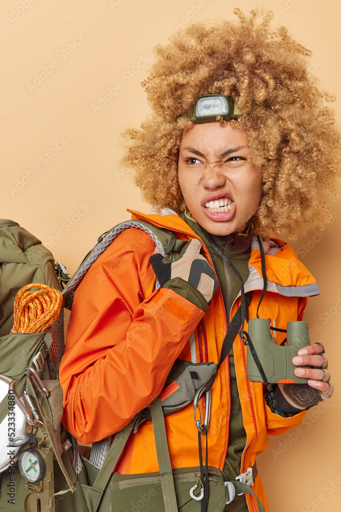 Angry irritated woman clenches teeth and frowns face holds binoculars carries heavy rucksack explores something during expedition wears headlamp and orange jacket isoated over beige background Angry irritated woman clenches teeth and frowns face holds binoculars carries heavy rucksack explores something during expedition wears headlamp and orange jacket isoated over beige background