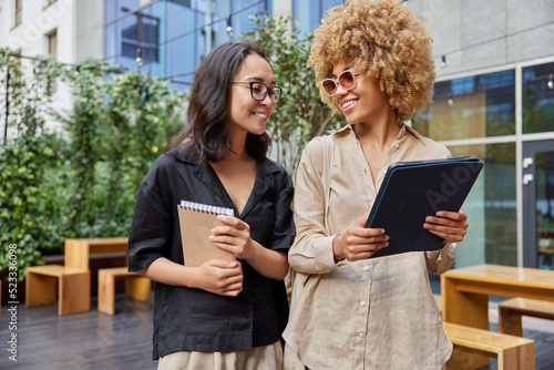 Multiracial young colleagues discuss business project while strolling outdoors against street cafe carry digital tablet and spiral notebook for writing notes dressed casually have glad expressions