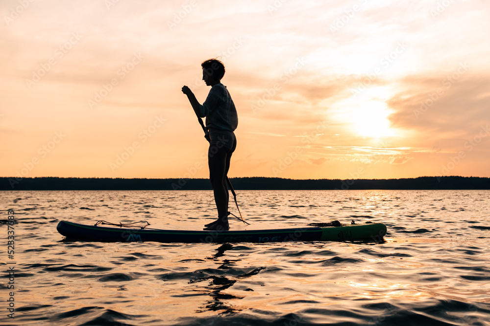 Joyful sport woman is training on a SUP board on a large lake during the evening. Sunset. Stand up paddle boarding - awesome active recreation in nature.