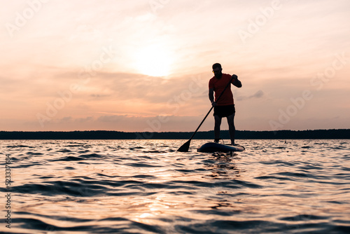 Joyful red bearded man in sunglasses is training on a SUP board on a large lake during sunny day. Stand up paddle boarding - awesome active recreation in nature. 