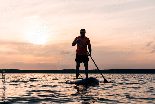 Joyful red bearded man in sunglasses is training on a SUP board on a large lake during sunny day. Stand up paddle boarding - awesome active recreation in nature. 