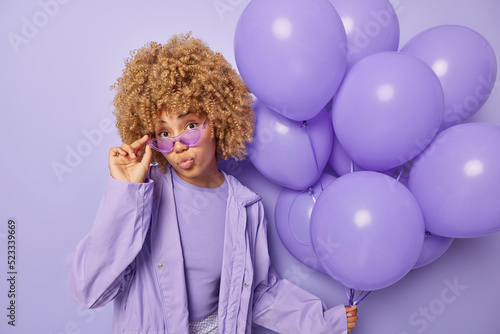 Stylish teenage girl wears sunglasses and windbreaker keeps lips folded comes on party to celebrate something holds bunch of inflated balloons isolated over purple background. Holiday concept