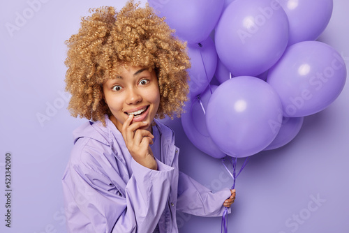 Surprised cheerful woman with curly blonde hair bites finger looks gladfully at camera wears jacket holds bunch of inflated balloons celebrates special occasion isolated over purple background