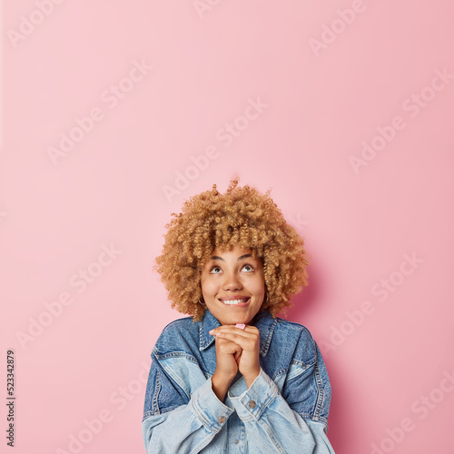 Dreamy curly haired European woman bites lips keeps hands under chin focused above dressed in denim jacket thinks about something isolated over pink background empty space for your promotion