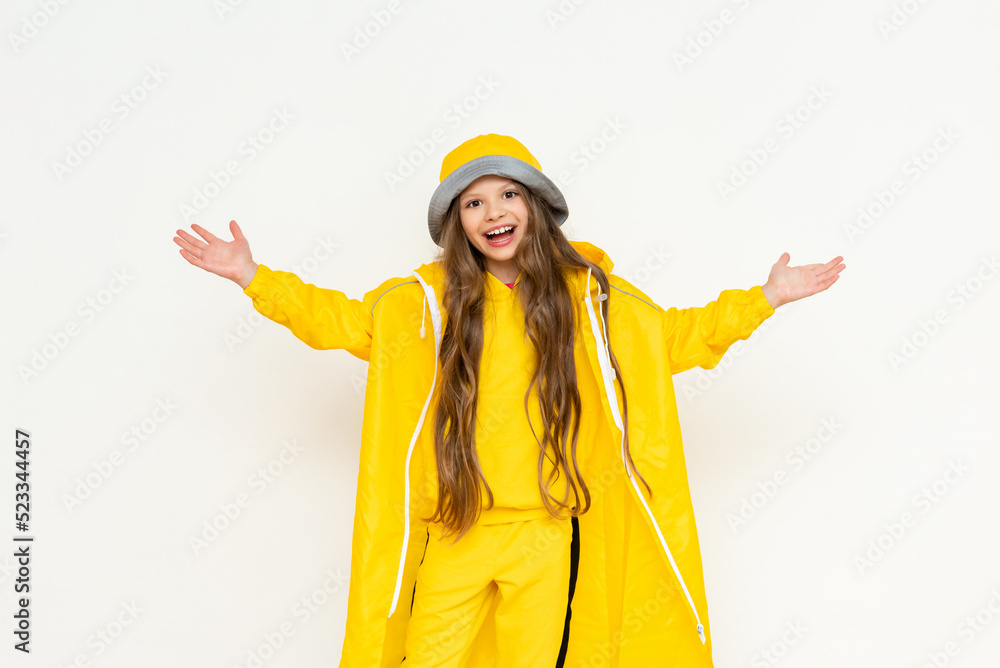 A little girl in a raincoat has spread her arms in different directions and is smiling broadly. A child in a yellow raincoat and panama hat on a white isolated background. A little girl in a raincoat has spread her arms in different directions and is smiling broadly. A child in a yellow raincoat and panama hat on a white isolated background.
