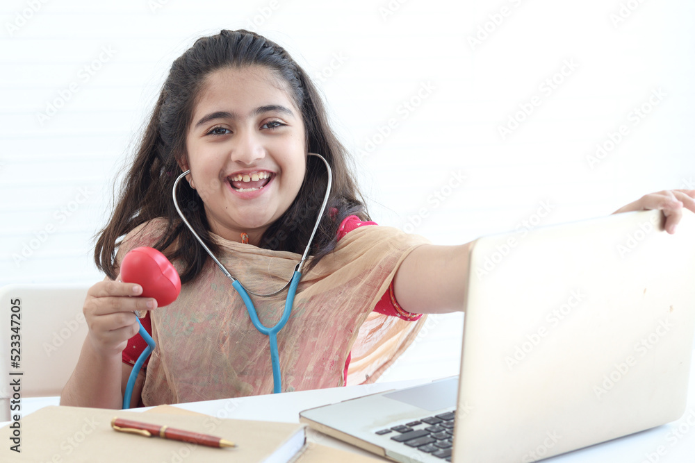 Portrait of adorable smiling Pakistani girl with beautiful eyes pretending to be doctor, Asian girl playing funny and holding doctor stethoscope, student kid using laptop computer on white background. Portrait of adorable smiling Pakistani girl with beautiful eyes pretending to be doctor, Asian girl playing funny and holding doctor stethoscope, student kid using laptop computer on white background.