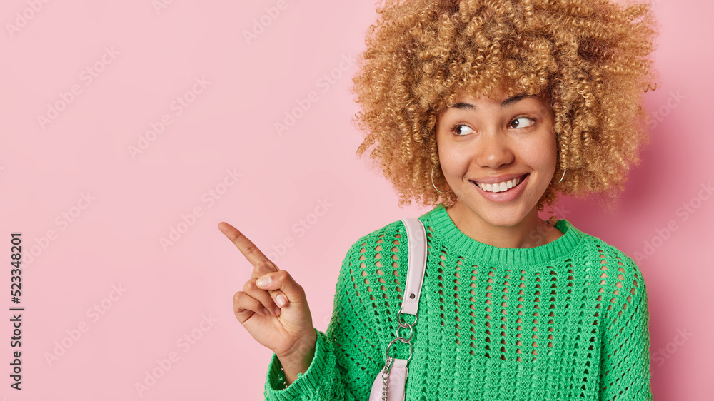 Check this out. Cheerful young woman with curly blonde hair dressed in green knitted jumper points index finger aside on blank space shows advertisement or promo isolated over pink background