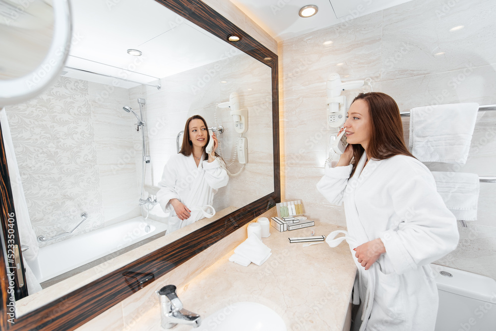 Fototapeta premium A young beautiful girl in a beautiful white bathroom talking on the phone. A fresh good morning at the hotel. Rest and travel. Hotel recreation, and tourism.