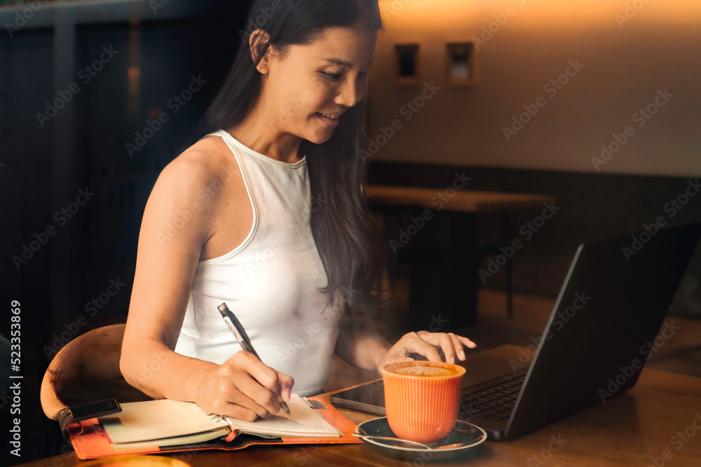 Smiling business woman writing and using laptop in coffee shop