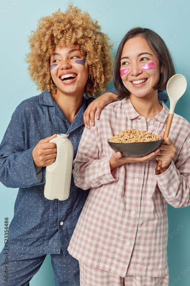 Vertical shot of diverse women smile broadly dressed in nightwear eat healthy delicious cornflakes apply hydrogel patches under eyes to reduce wrinkles or puffiness isolated over blue background Vertical shot of diverse women smile broadly dressed in nightwear eat healthy delicious cornflakes apply hydrogel patches under eyes to reduce wrinkles or puffiness isolated over blue background