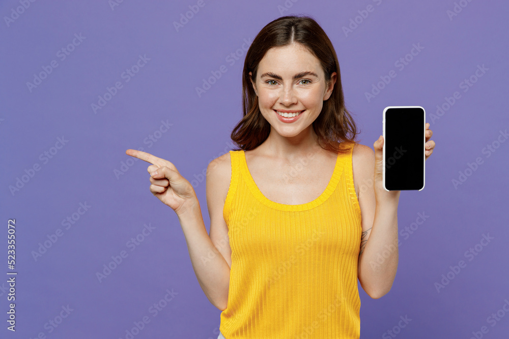 Young happy smiling fun woman 20s wear yellow tank shirt hold in hand use mobile cell phone with blank screen workspace area point index finger aside isolated on plain pastel light purple background. Young happy smiling fun woman 20s wear yellow tank shirt hold in hand use mobile cell phone with blank screen workspace area point index finger aside isolated on plain pastel light purple background.