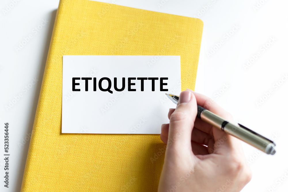 A white business card with ETIQUETTE text on a billboard on a white background next to the hand of a girl with a pen writes text A white business card with ETIQUETTE text on a billboard on a white background next to the hand of a girl with a pen writes text