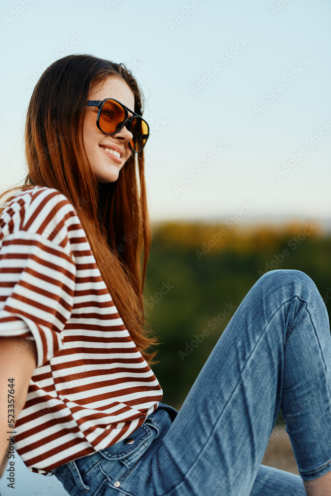A fashion woman in stylish sunglasses, a striped t-shirt and jeans sits on the trunk of a car and looks at the beautiful nature of autumn. Travel lifestyle A fashion woman in stylish sunglasses, a striped t-shirt and jeans sits on the trunk of a car and looks at the beautiful nature of autumn. Travel lifestyle