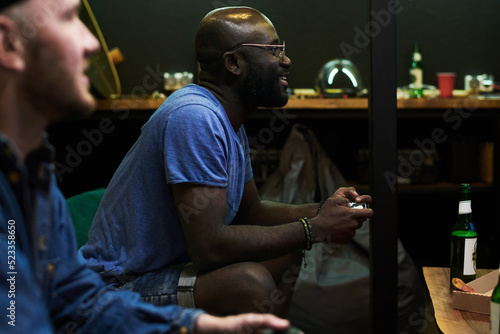 Happy young African American man in blue t-shirt playing video game while sitting on couch and pressing buttons on gamepad
