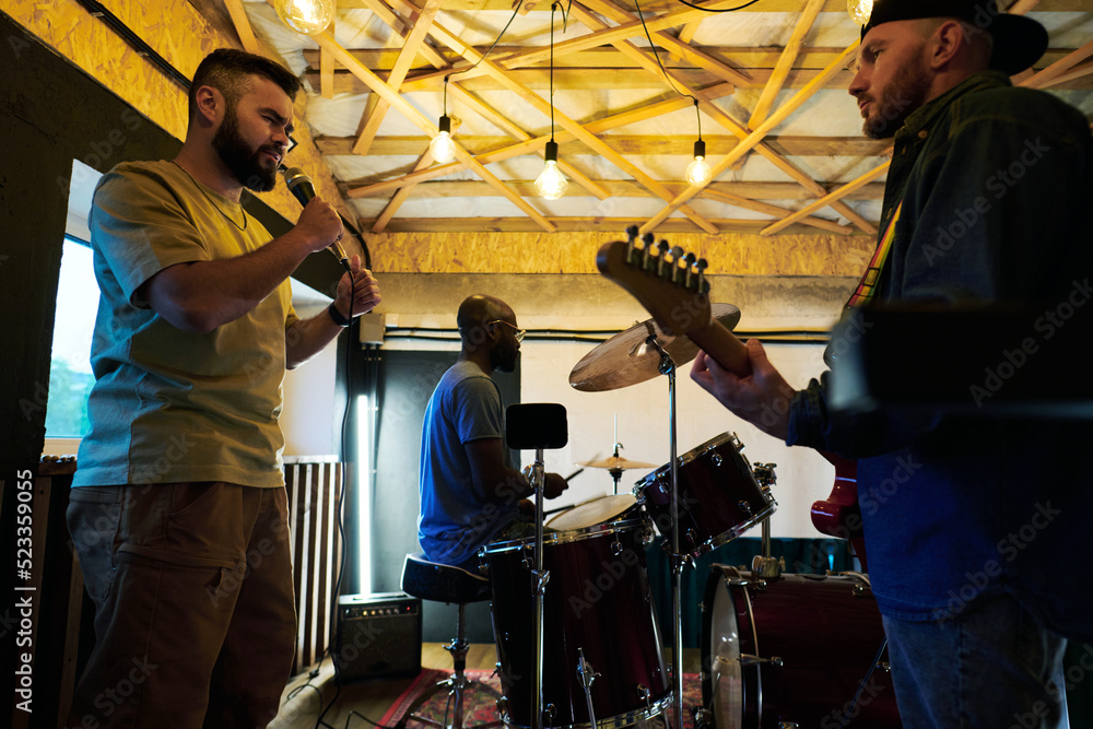Bearded man singing in microphone among his buddies playing electric guitar and hitting drum set during repetition in garage