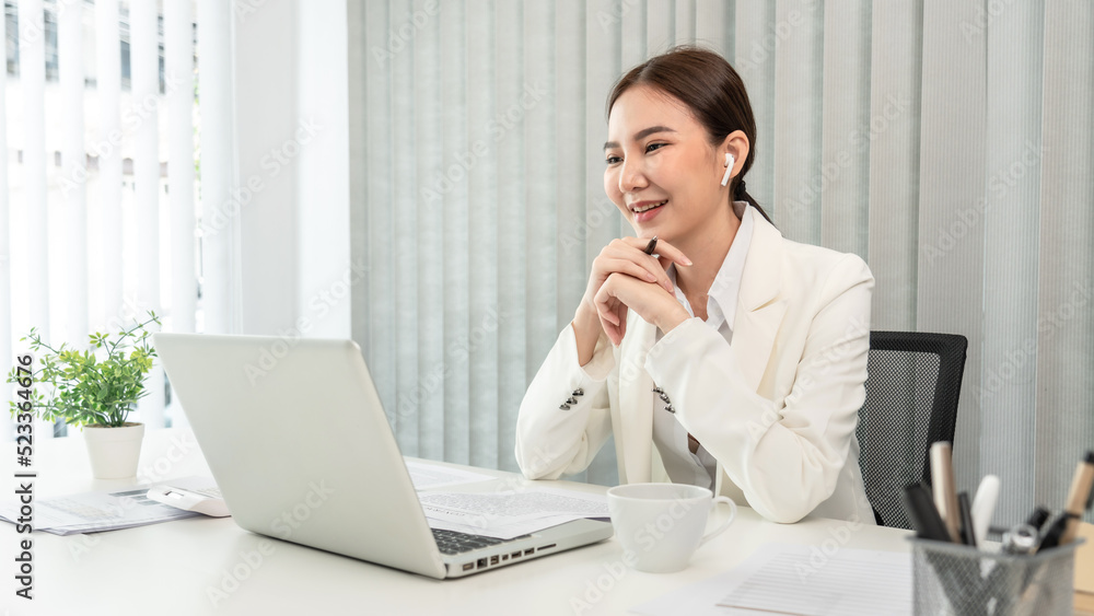 Asian businesswoman talking on the wireless headphones with a colleague in a private office, Telephone communication, Emote conversation, Modern office with laptop and documents on the desk Asian businesswoman talking on the wireless headphones with a colleague in a private office, Telephone communication, Emote conversation, Modern office with laptop and documents on the desk