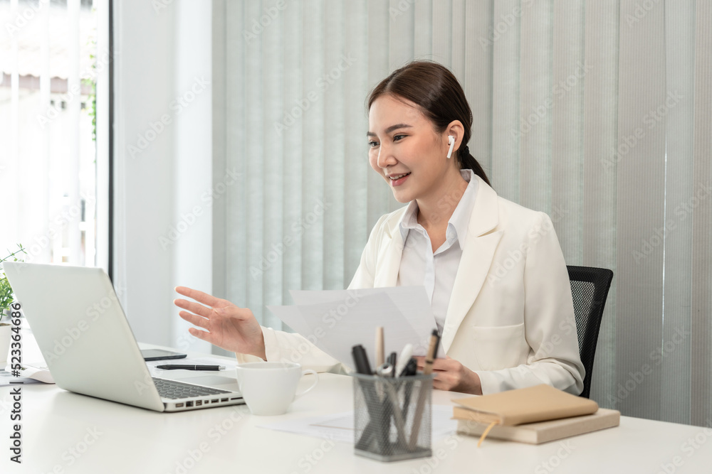 Asian businesswoman talking on the wireless headphones with a colleague in a private office, Telephone communication, Emote conversation, Modern office with laptop and documents on the desk Asian businesswoman talking on the wireless headphones with a colleague in a private office, Telephone communication, Emote conversation, Modern office with laptop and documents on the desk