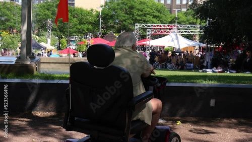 Short movie shot from the back of an elderly man, sitting in mobility scooter watching a crowd of people gathered for a summer fair in a Montreal park.