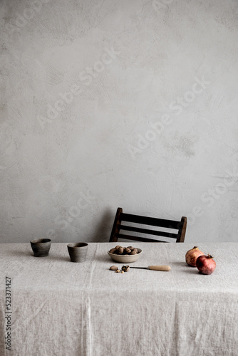 Autumn set up inside with wooden chair and table covered with beige linen tablecloth, coffee in ceramic grey cups, pomegranade, and walnut against a textured grey wall as background.  