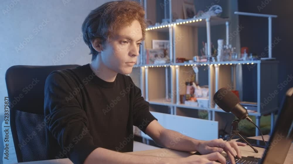 The blogger is preparing for the broadcast, writes text on a laptop, there is a microphone on the table nearby. dark-haired guy. Vlogger, streamer. Young man.Handsome guy. The blogger is preparing for the broadcast, writes text on a laptop, there is a microphone on the table nearby. dark-haired guy. Vlogger, streamer. Young man.Handsome guy.