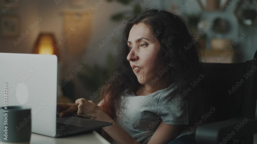 Woman with a disability sitting in a wheelchair at the table in a living room, and browsing the internet or social media swiping on laptop touchpad Woman with a disability sitting in a wheelchair at the table in a living room, and browsing the internet or social media swiping on laptop touchpad