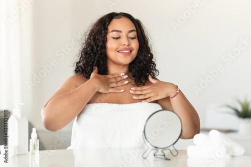 Young black plus size woman touching decollete zone and looking in mirror, enjoying domestic beauty treatments photo