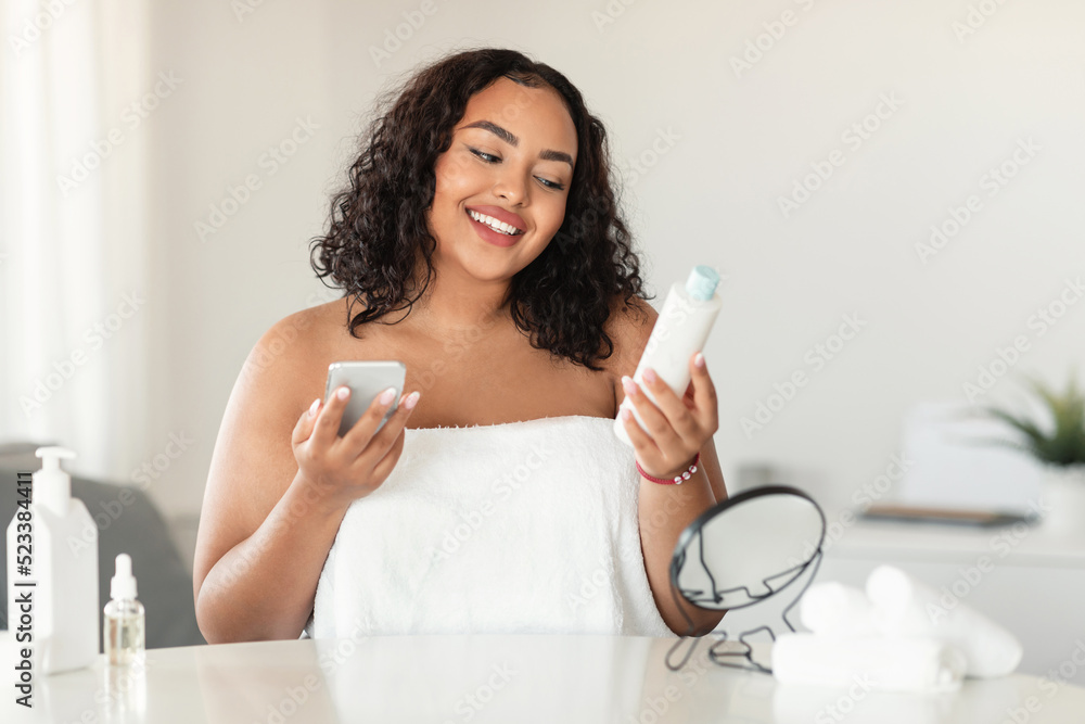 Happy black chubby woman in white towel reading recommendations on lotion bottle and using smartphone Happy black chubby woman in white towel reading recommendations on lotion bottle and using smartphone