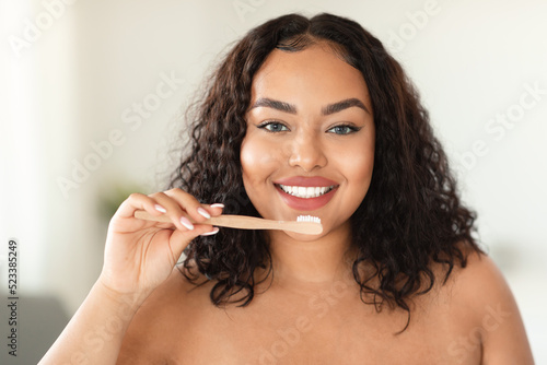 Happy black chubby woman standing in bathroom  brushing her teeth in the morning  looking at camera