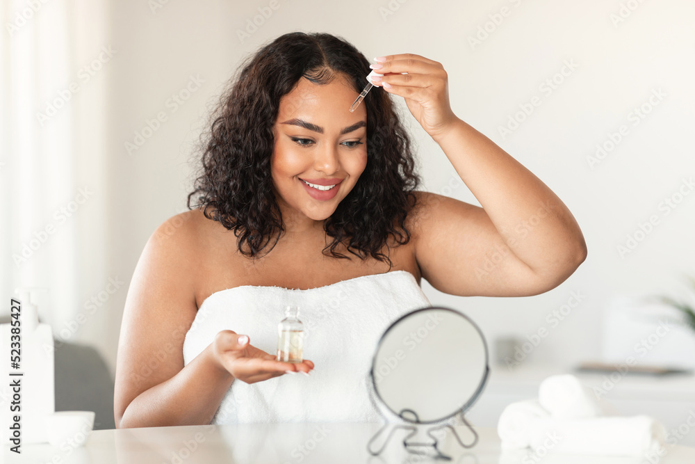 Skincare concept. Pretty black chubby woman applying face serum with dropper, sitting in front of mirror and smiling Skincare concept. Pretty black chubby woman applying face serum with dropper, sitting in front of mirror and smiling