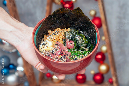 man's hand is holding a poke salad with tuna - a traditional dish of Hawaiian and Japanese cuisine. Christmas balls are visible in the background.