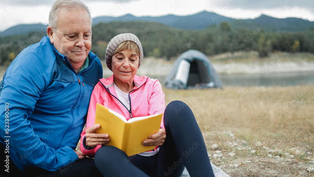 Happy senior couple having leisure time reading a book camping in mountain lake - Focus on man face