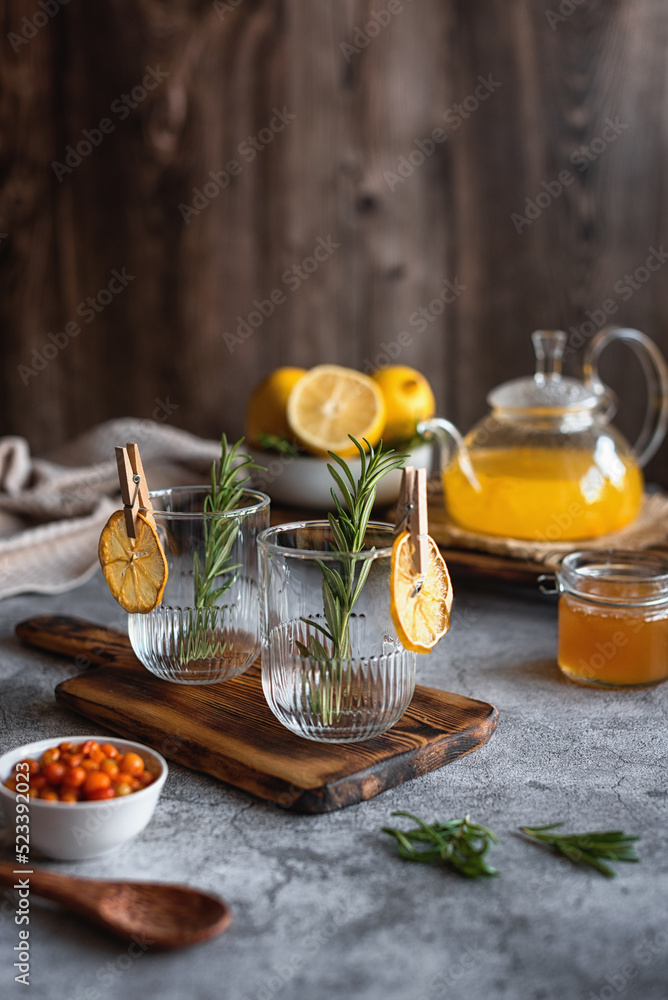 Empty glass mugs decorated with rosemary for serving sea buckthorn tea. A warming winter drink. Vitamin tea. Sea buckthorn tea with honey, lemon and ginger on a gray background. Empty glass mugs decorated with rosemary for serving sea buckthorn tea. A warming winter drink. Vitamin tea. Sea buckthorn tea with honey, lemon and ginger on a gray background.